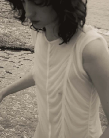 Woman in white dress on a rocky beach with waves in the background, creating a serene and muted coastal scene. Fashion photography.