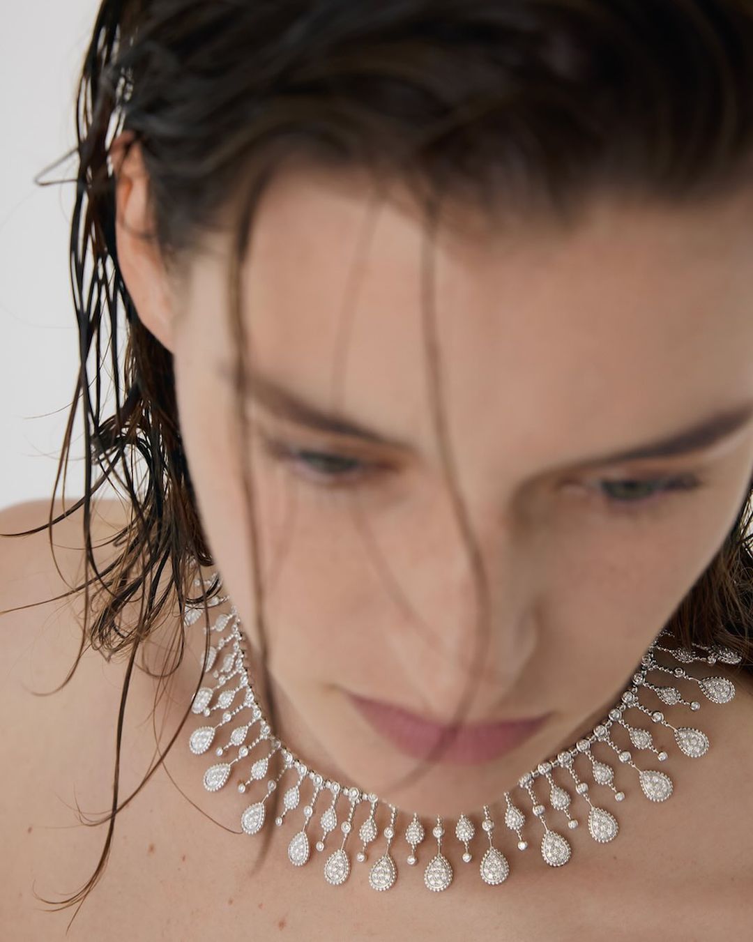 Close-up of an adult female with wet hair, fair skin, and an elaborate diamond necklace looking downward in a neutral studio.