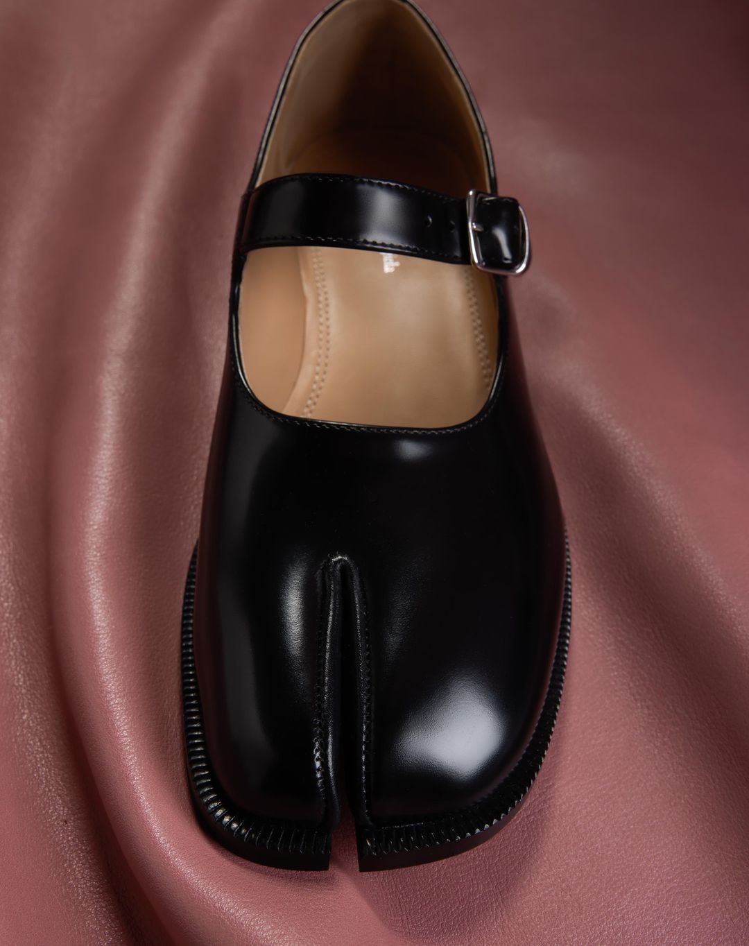 Close-up studio shot of a glossy black Tabi Mary Jane shoe with a buckled strap set against a softly draped pink background.