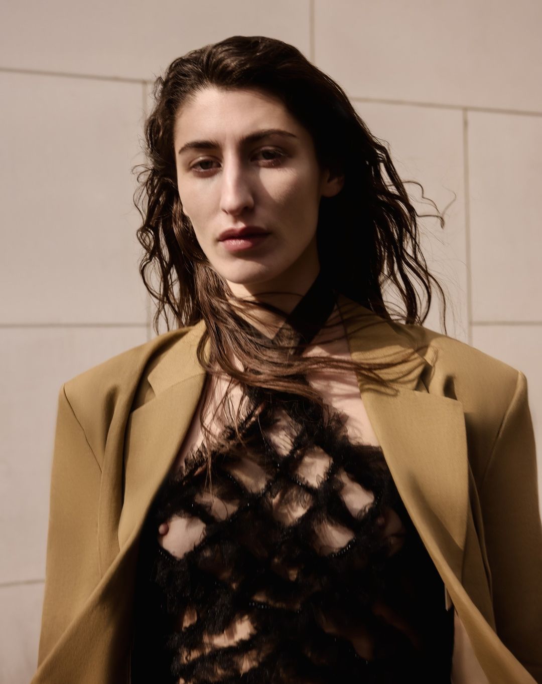 Portrait of a dark-haired woman wearing a tan jacket and black sheer top in an outdoor setting, looking directly at the camera.