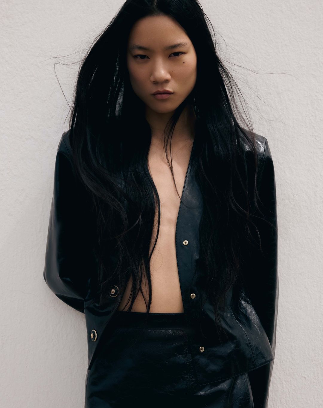 Portrait of a woman with long black hair wearing a black leather jacket and skirt, posing against a white background in a fashion editorial.