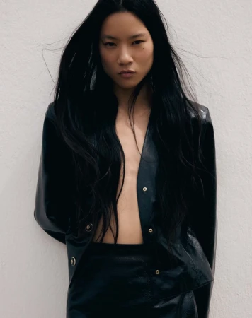 Portrait of a woman with long black hair wearing a black leather jacket and skirt, posing against a white background in a fashion editorial.