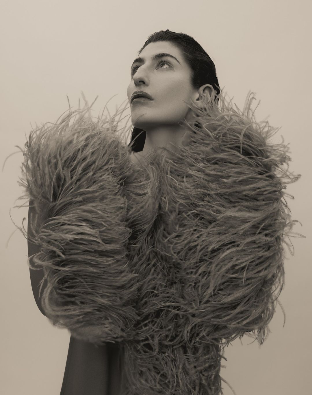 Portrait of a woman with feathered garment looking up in a studio setting, featuring neutral tones and editorial style.