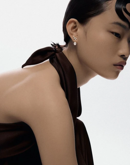 Close-up of Asian woman with bare shoulder and brown fabric, showcasing small elegant earrings in a brightly lit studio.