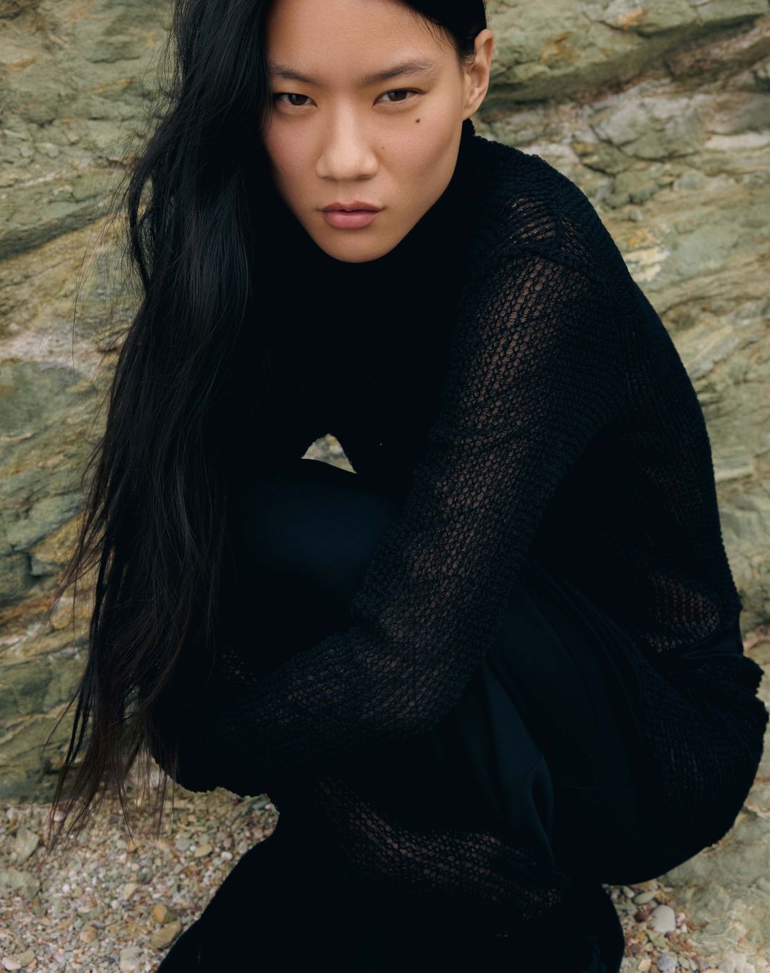 Adult female fashion model with long black hair wearing a black outfit crouching outdoors against a rock wall looking at the camera.