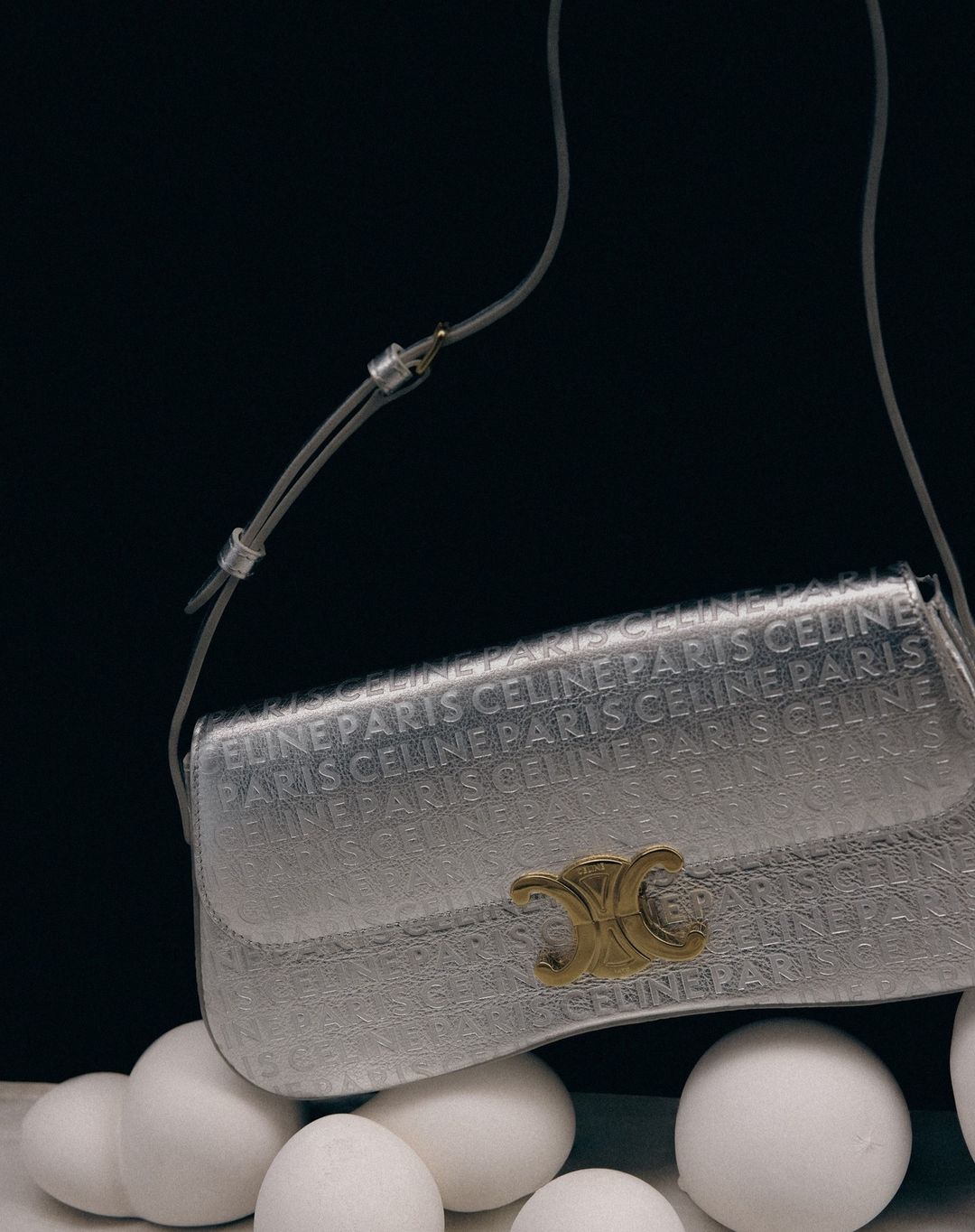 Celine Paris handbag in silver with logo pattern displayed on a bed of white spheres against a dark background, studio lighting.