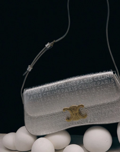 Celine Paris handbag in silver with logo pattern displayed on a bed of white spheres against a dark background, studio lighting.