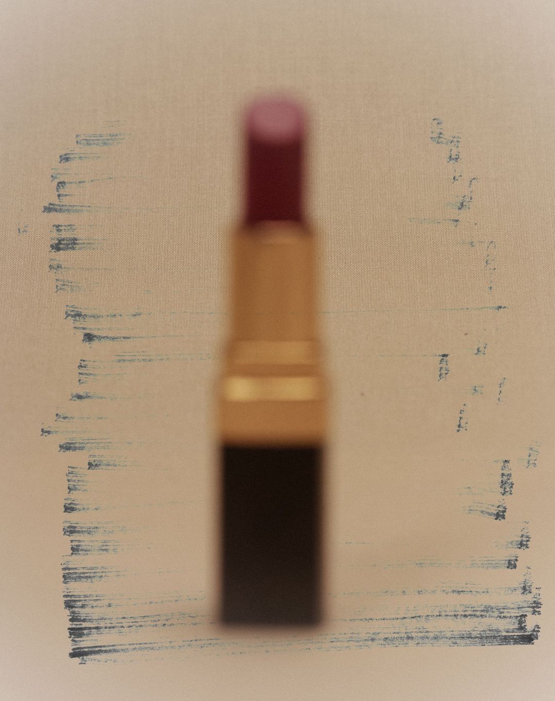 Close-up of blurred lipstick with a gold tube and red pigment on a beige background with faded gray brush stroke accents.