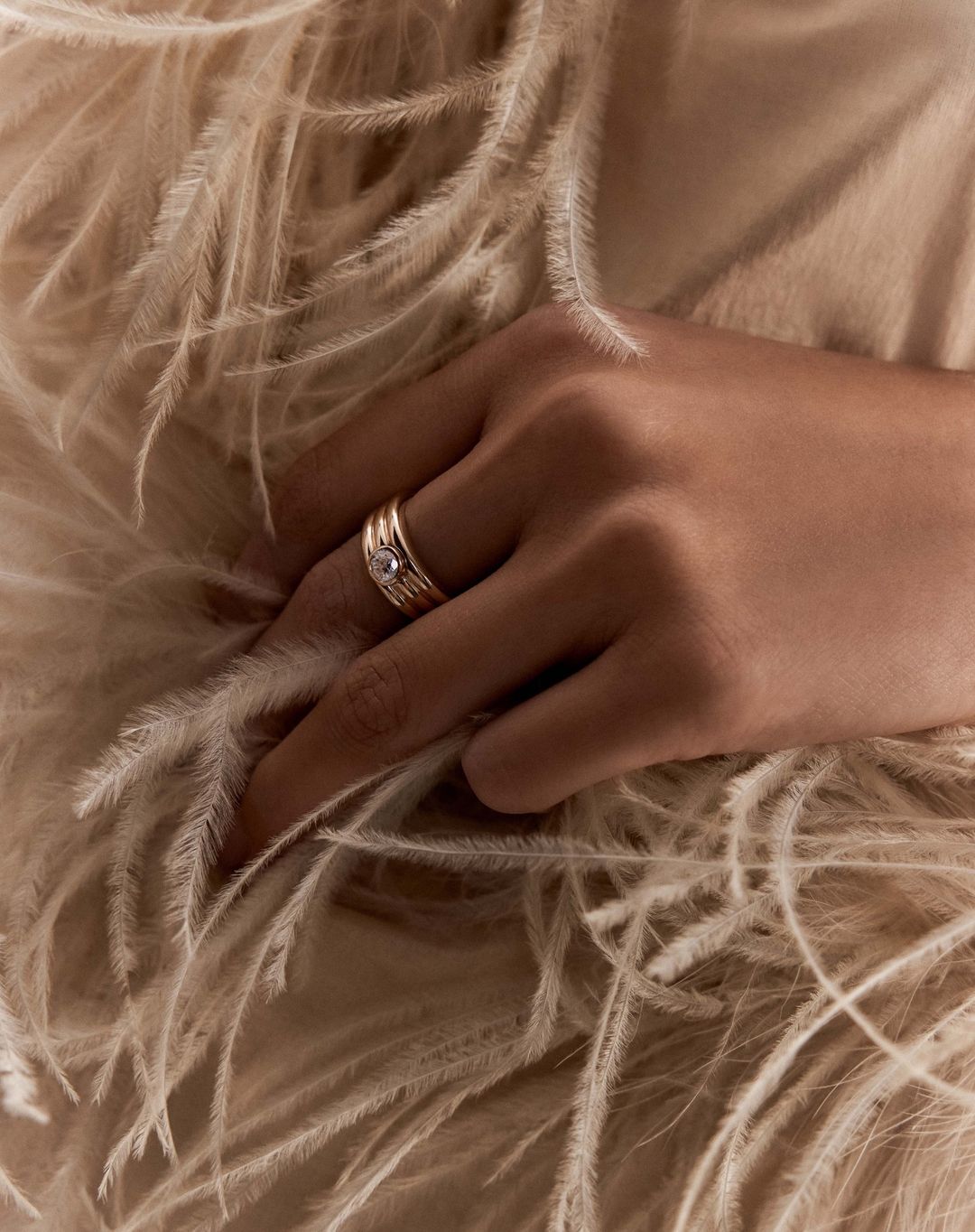 Close-up of a person's hand adorned with gold rings, resting against a background of soft, cream-colored feathers.
