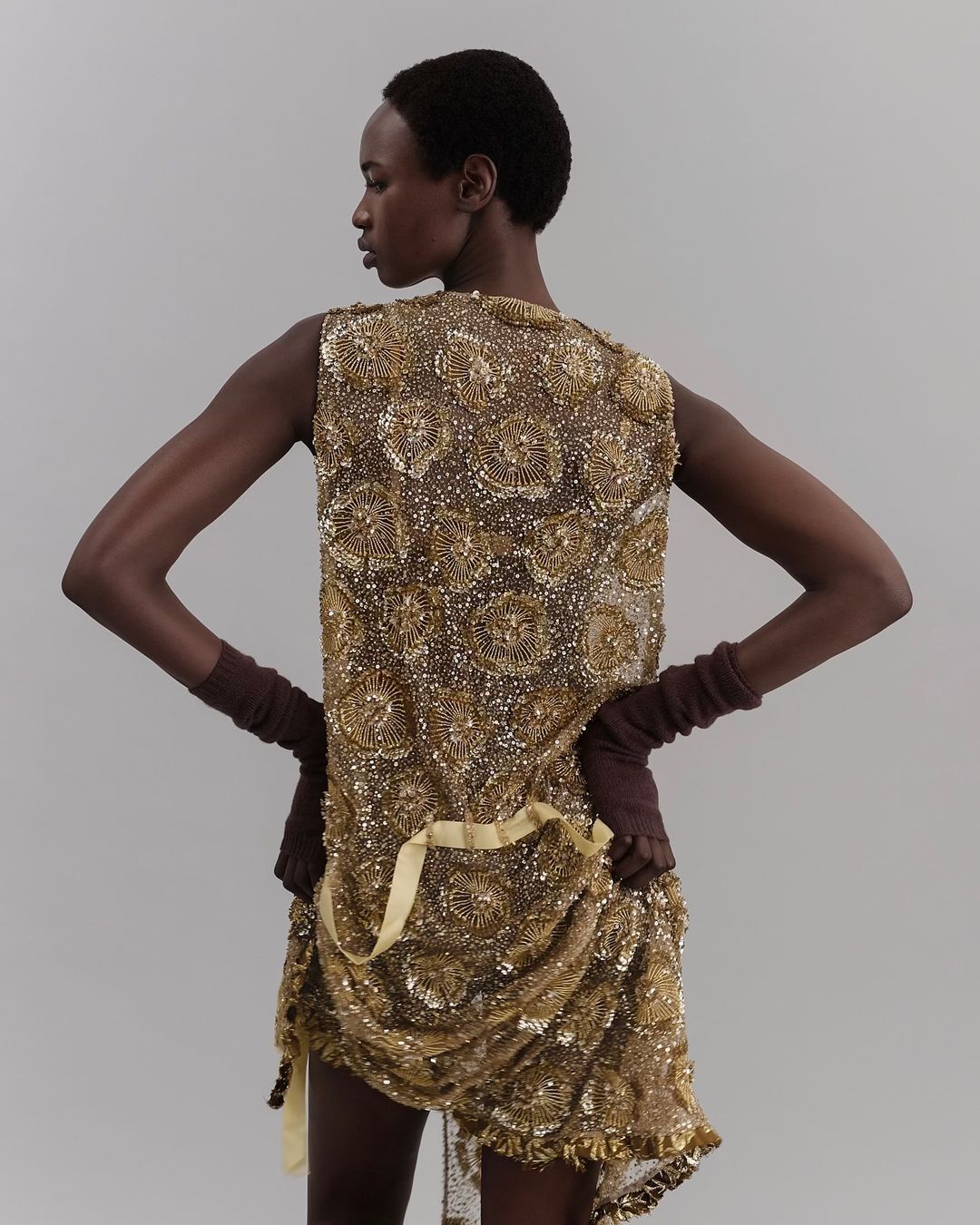 Woman in golden beaded dress with fingerless gloves poses against a plain background, showcasing fashion and design.