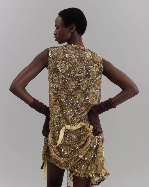Woman in golden beaded dress with fingerless gloves poses against a plain background, showcasing fashion and design.