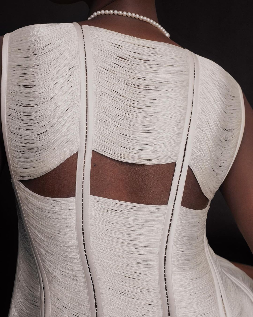 Back view of woman wearing white dress with layered fringe design and pearl necklace against a dark background in studio lighting.