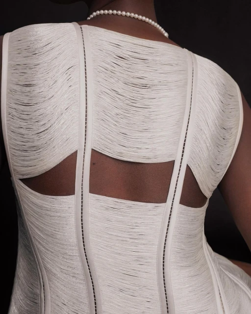 Back view of woman wearing white dress with layered fringe design and pearl necklace against a dark background in studio lighting.