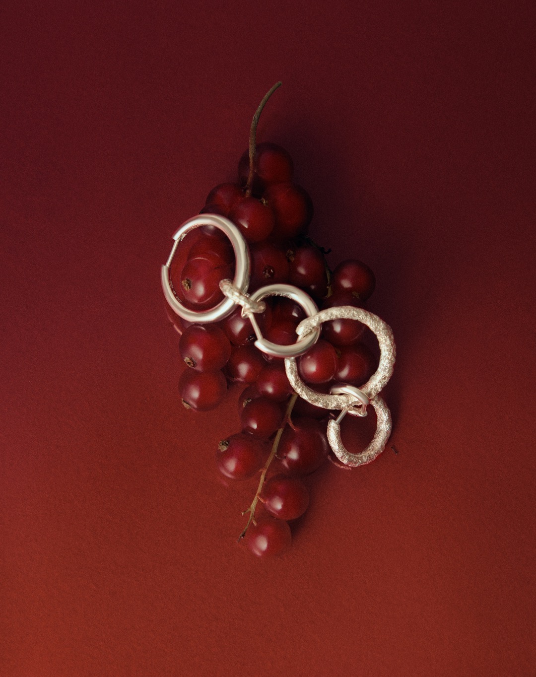 Still life studio shot of linked silver earrings with red currant berries on a red gradient backdrop, high-angle product photo.