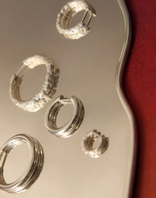 Close-up studio shot of four elegant silver hoop earrings, with varied textures, placed on a reflective surface with red background accent.