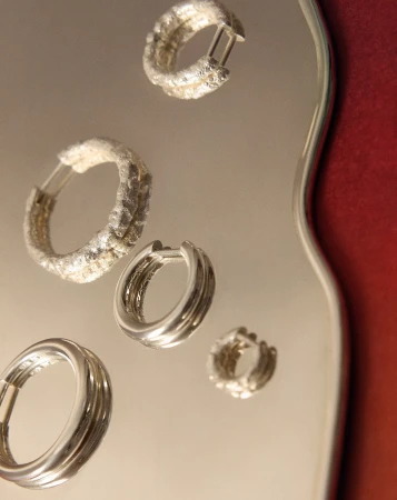 Close-up studio shot of four elegant silver hoop earrings, with varied textures, placed on a reflective surface with red background accent.
