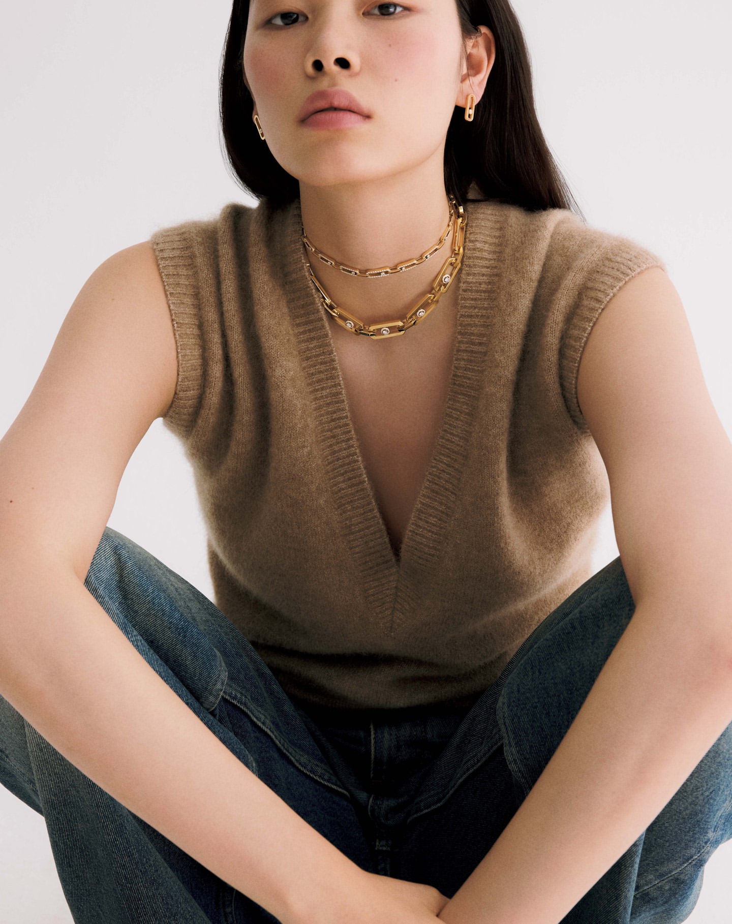 Woman wearing a brown sleeveless V-neck knit sweater and layered gold necklaces against a plain white background