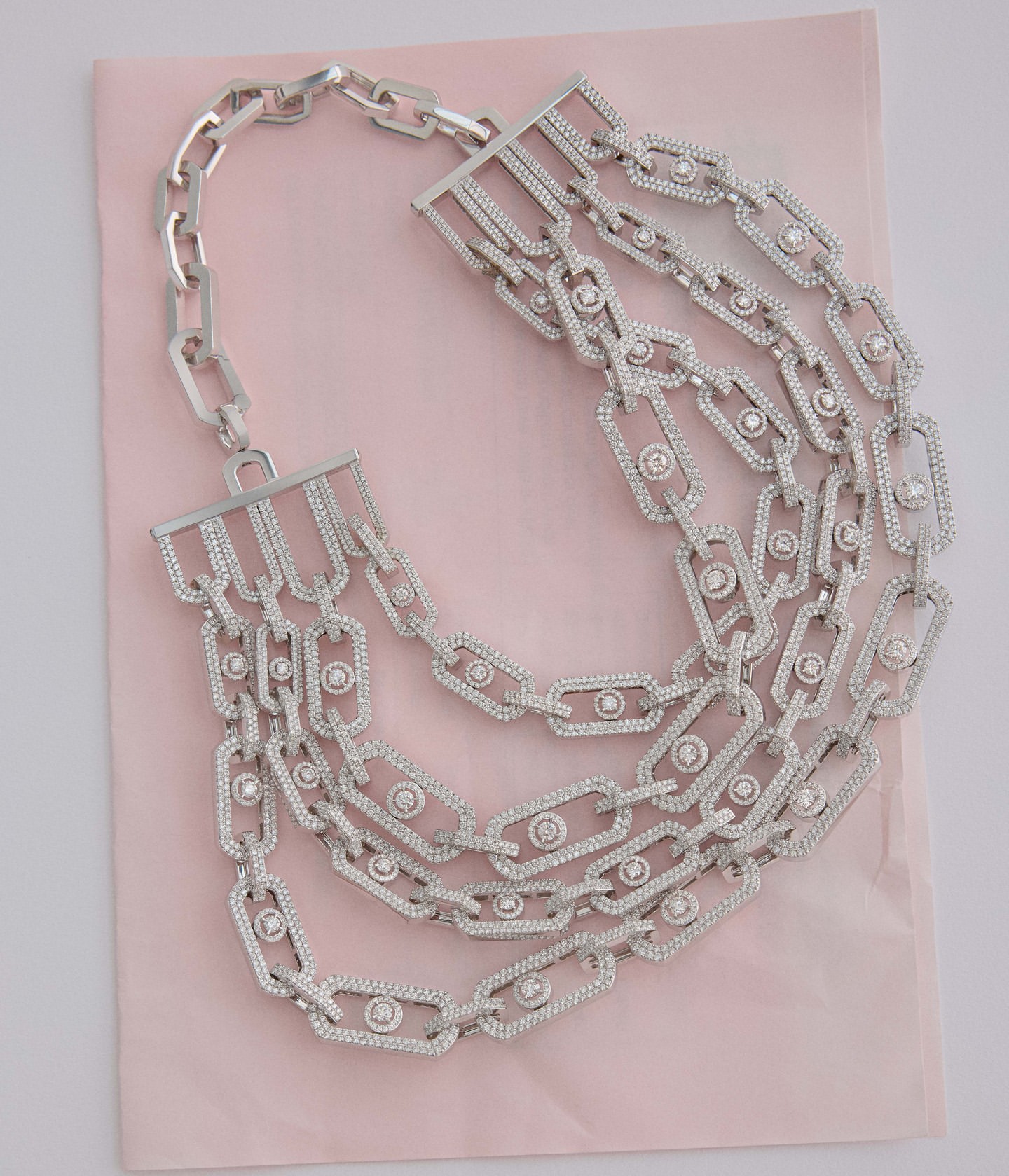 A close-up studio shot of a multi-strand silver chain necklace with sparkling stone details, laying on a light pink surface. Jewelry.