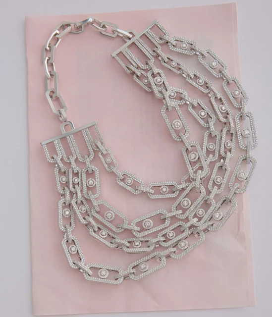 A close-up studio shot of a multi-strand silver chain necklace with sparkling stone details, laying on a light pink surface. Jewelry.