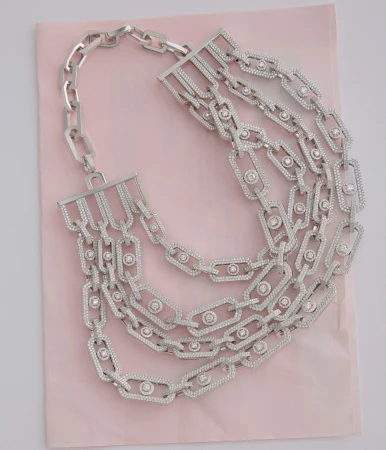 A close-up studio shot of a multi-strand silver chain necklace with sparkling stone details, laying on a light pink surface. Jewelry.