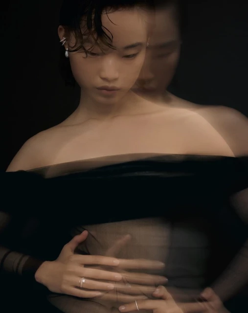 Asian woman in off-the-shoulder black top poses in a blurred studio shot with dark backdrop, showcasing rings and earrings.