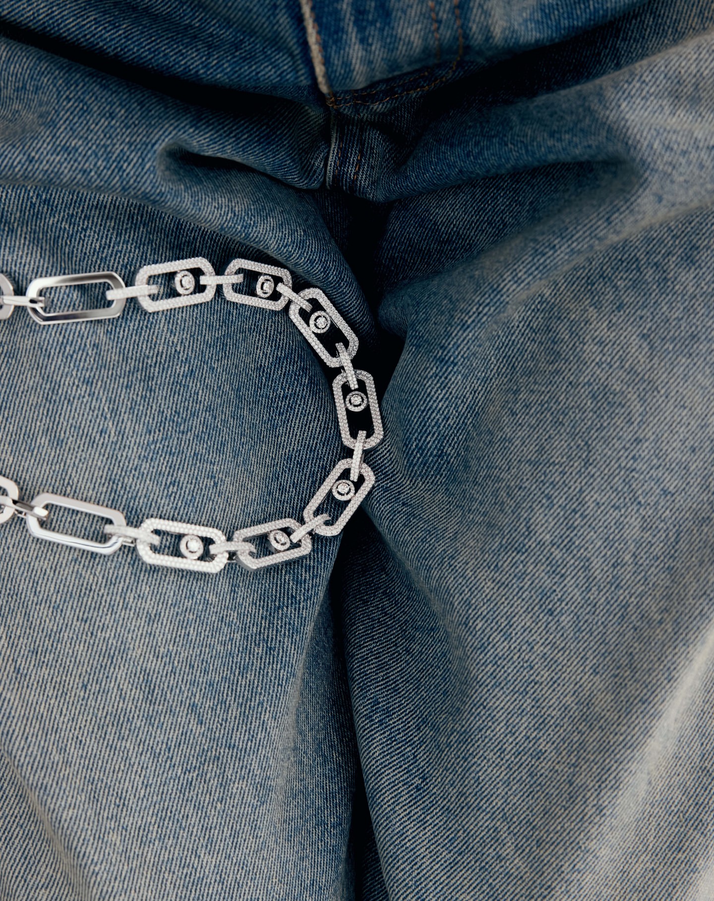 A close-up of a silver chain necklace with circular accents, draped over a pair of light blue denim jeans.