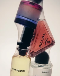 Close-up of stacked fragrance bottles including Prada and Louis Vuitton on a seamless white background.