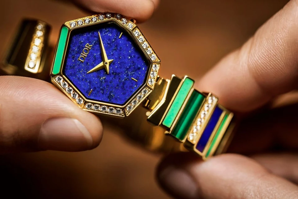Close-up of a gold and gemstone Dior watch held in someone's fingers, showcasing its blue face, green accents, and diamond detailing.