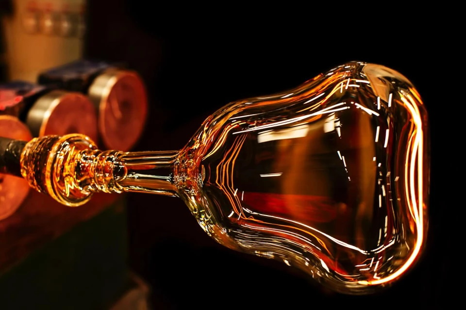 Close-up of a translucent, amber glass vessel, showcasing artistic glassblowing in a dark studio setting with tools in the background.