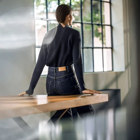 Model with braided hair wearing a dark ribbed sweater and dark denim jeans seated on a wooden table in an interior space