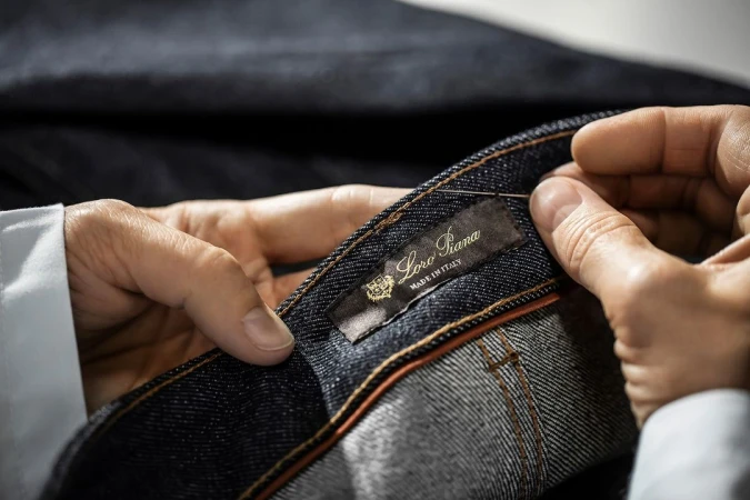 Close-up of hands sewing a Loro Piana brand label on dark blue denim jeans, highlighting craftsmanship and attention to detail in fashion.