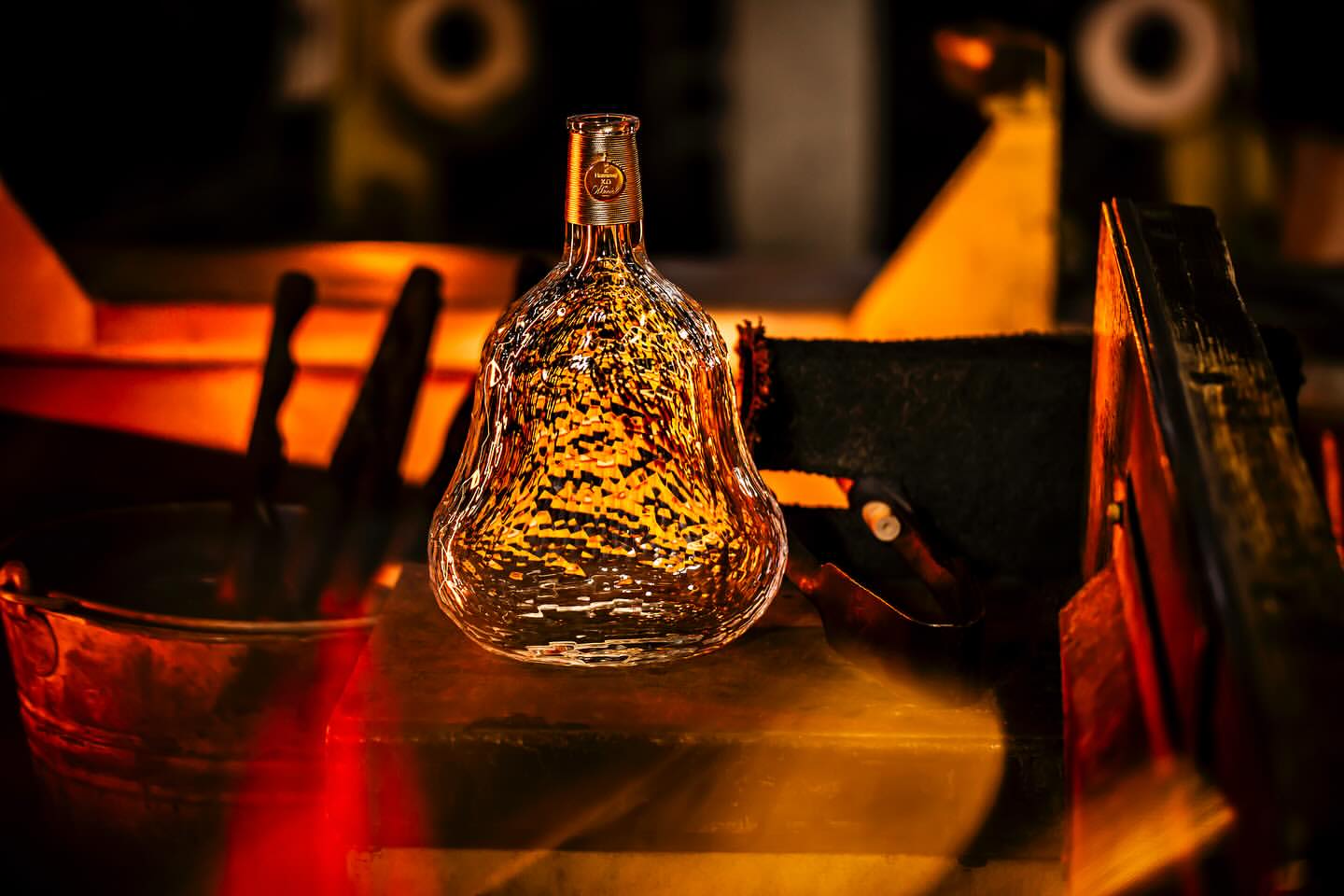 Clear glass liquor bottle with textured surface sits on a reflective surface with out-of-focus industrial background in warm light.