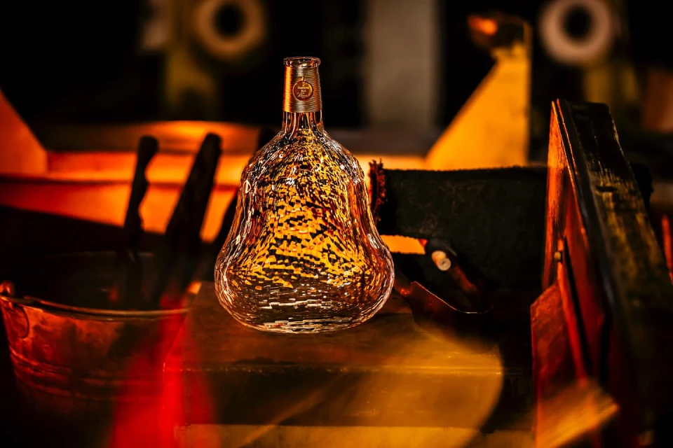 Clear glass liquor bottle with textured surface sits on a reflective surface with out-of-focus industrial background in warm light.