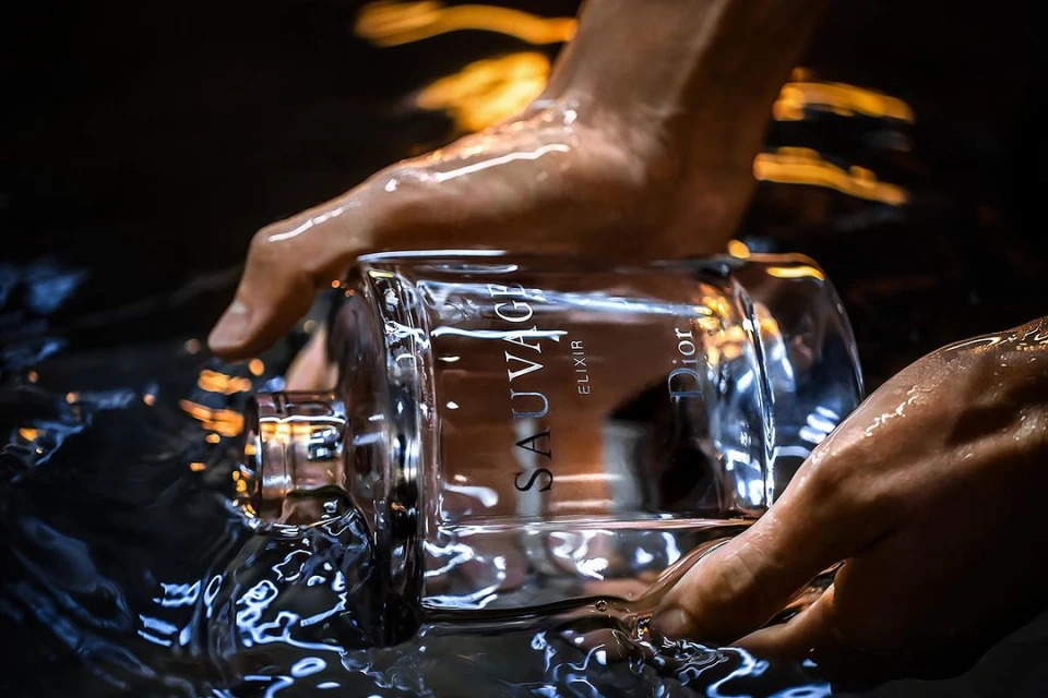 Close-up of Dior Sauvage Elixir bottle being held in hands with water splashing around it, product shot, fragrance advertisement.