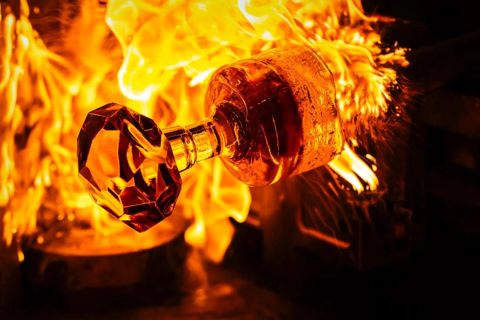 Close-up of a clear glass perfume bottle with a faceted stopper enveloped in bright orange flames in a dark, industrial setting.