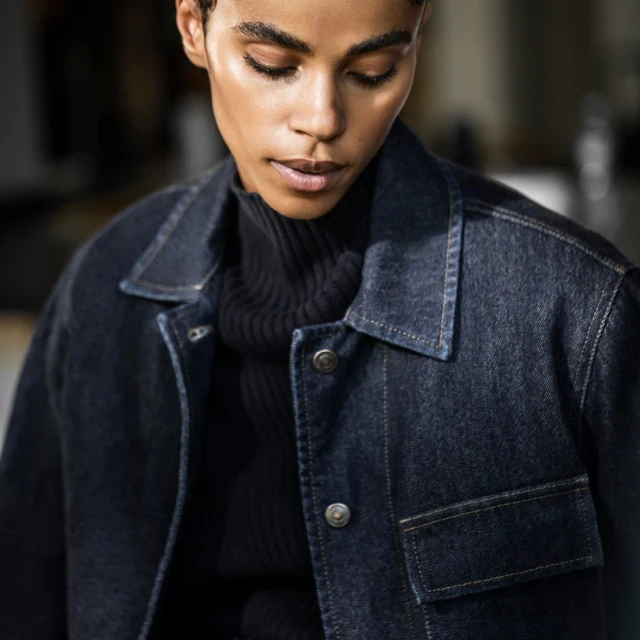 A fashionable adult female with short dark hair gazes downward while posing indoors wearing a denim jacket and a dark turtleneck sweater.