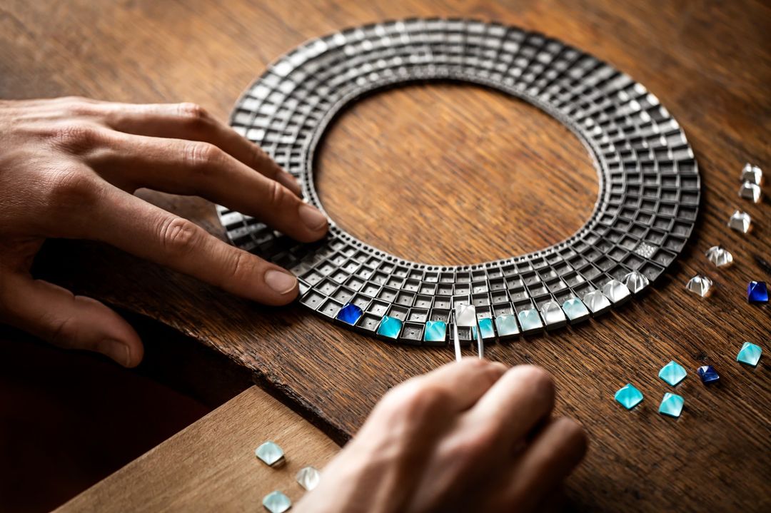 Craftsperson meticulously sets blue gems into a modern silver necklace on a wooden workbench in a close-up product shot.