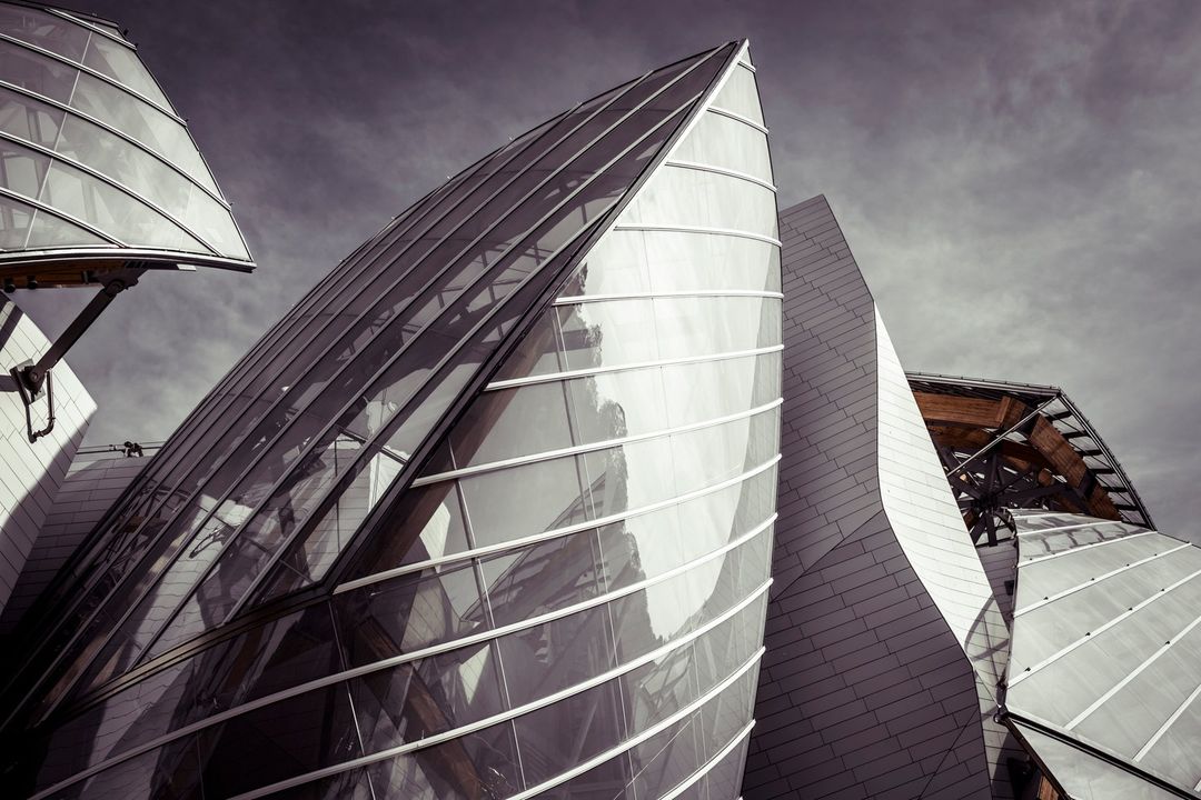 Architectural detail of the Fondation Louis Vuitton in Paris, featuring glass and grey stone against a cloudy sky, creating a modern, artistic design.