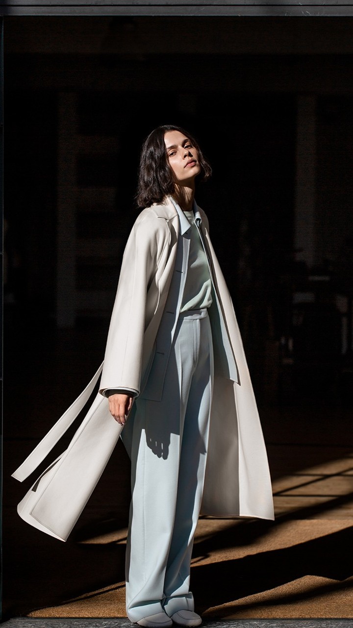 Fashion shot of a woman in a white coat and light blue pants standing in a doorway with light and shadow on the floor.