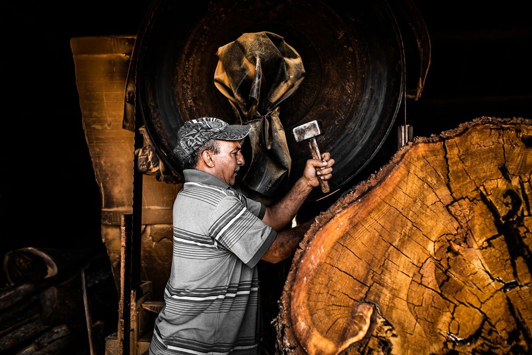 Adult male wearing a hat hammering a large wooden log next to a saw blade in a dimly lit indoor environment.