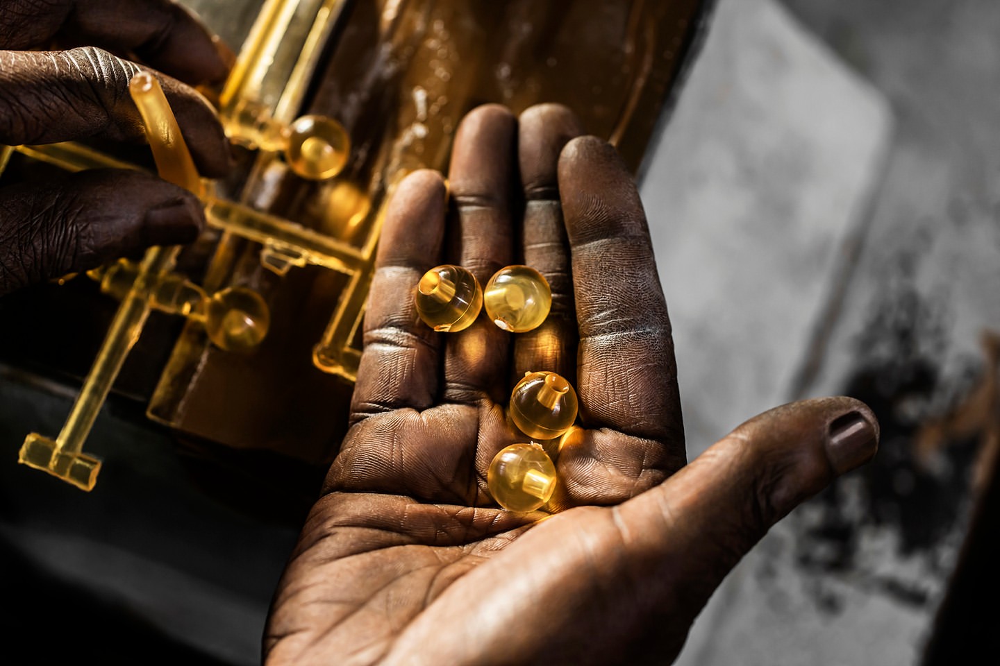 Close-up of dark-skinned hands holding five translucent yellow gel beads; a yellow translucent object is in the soft-focus background.