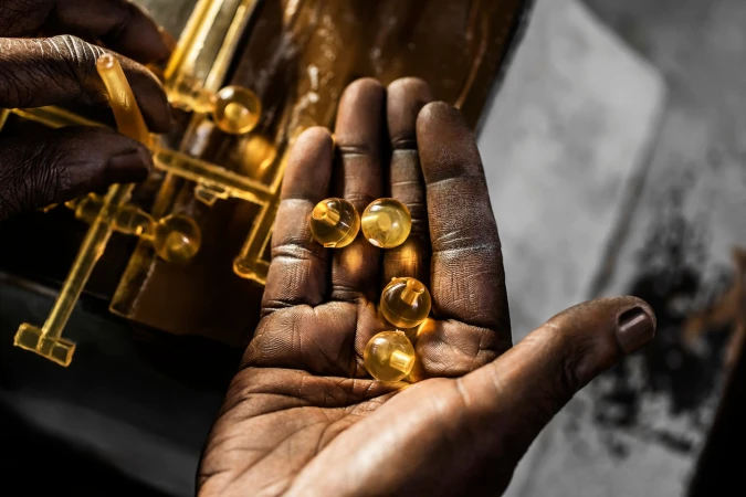 Close-up of dark-skinned hands holding five translucent yellow gel beads; a yellow translucent object is in the soft-focus background.