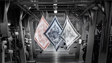 Three patterned scarves hang suspended within a high-ceilinged textile factory. Industrial equipment lines the floor in monochromatic tones.