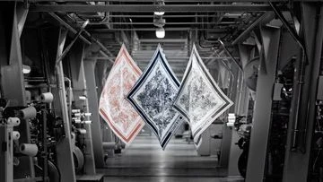 Three patterned scarves hang suspended within a high-ceilinged textile factory. Industrial equipment lines the floor in monochromatic tones.