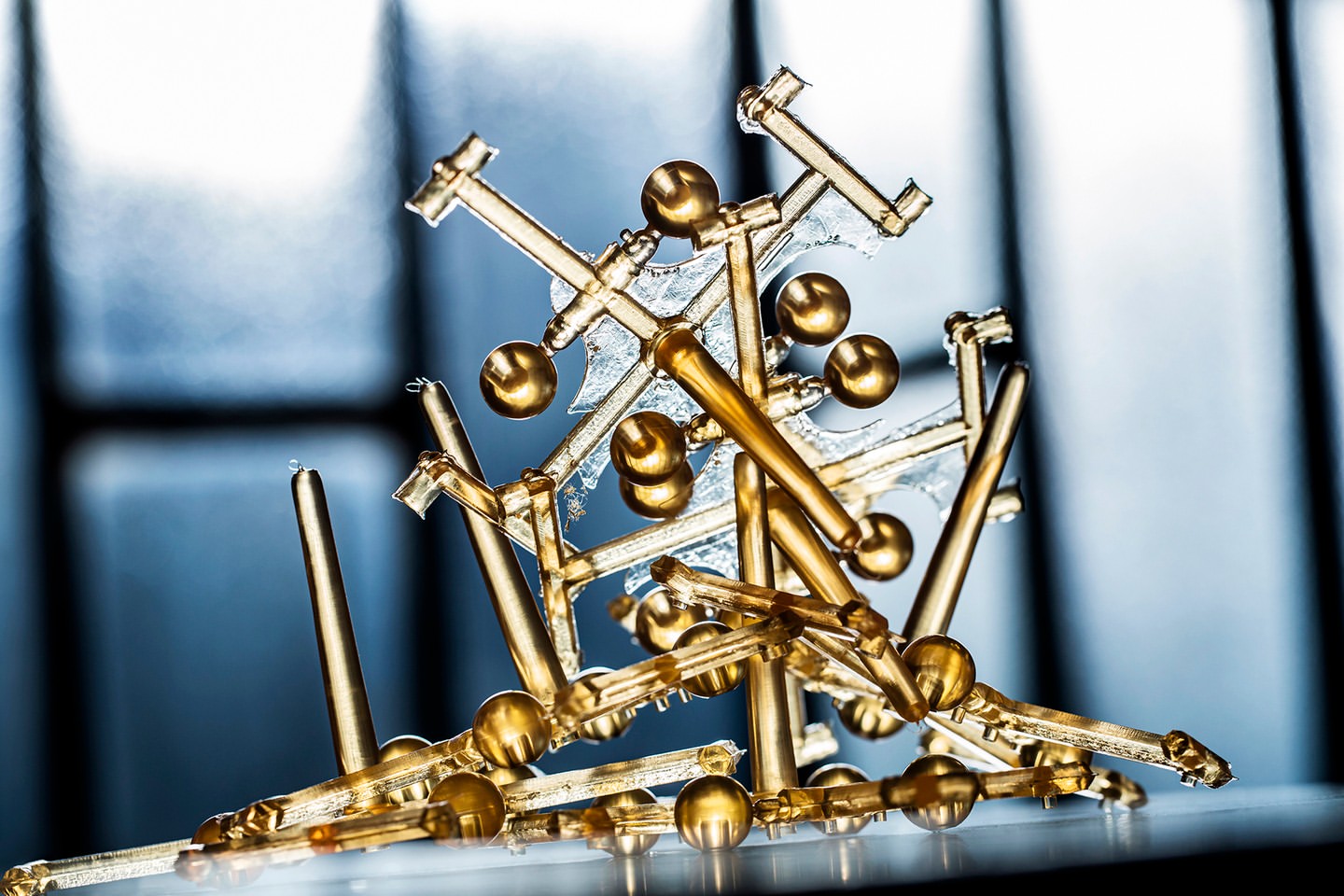 Close-up of gold-colored abstract objects, including rods and spheres, against a blurred blue and white background, with a shiny surface.
