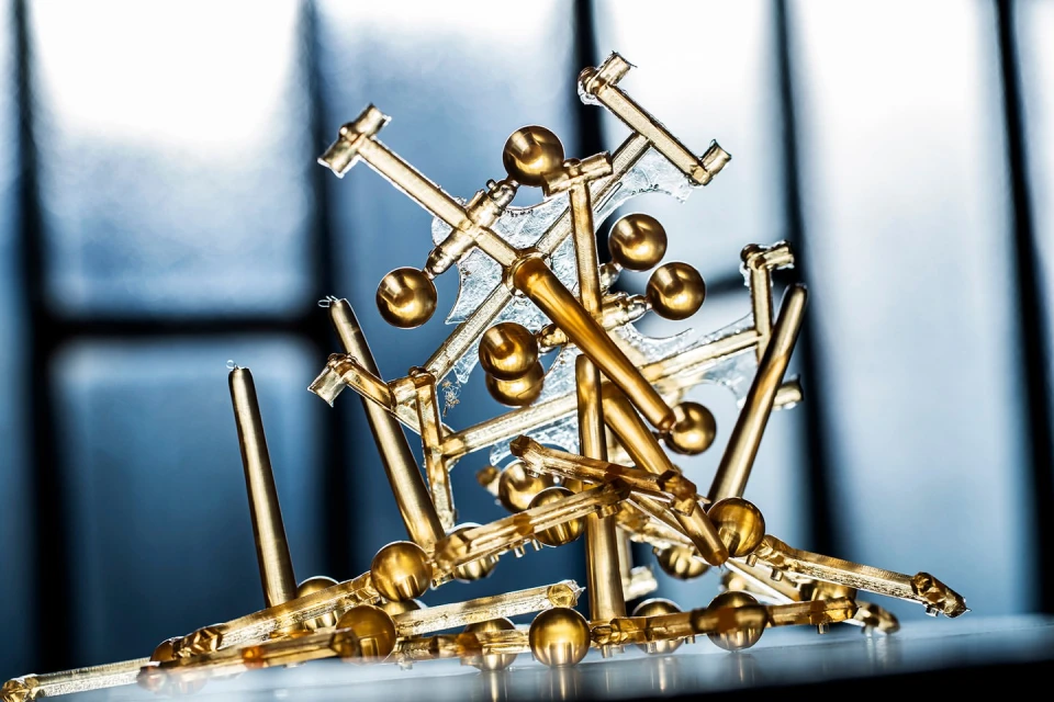Close-up of gold-colored abstract objects, including rods and spheres, against a blurred blue and white background, with a shiny surface.