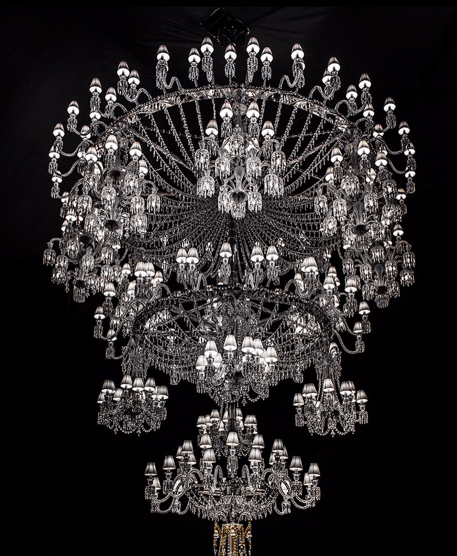 Extravagant multi-tiered crystal chandelier with dozens of light bulbs against a solid black background in an indoor setting.