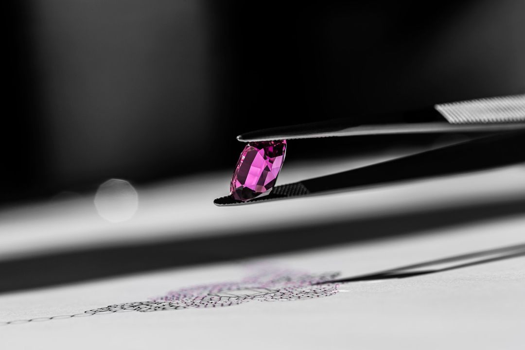 Close-up of pink gemstone held by metal tweezers, casting shadow on white surface against dark background with a circular light.