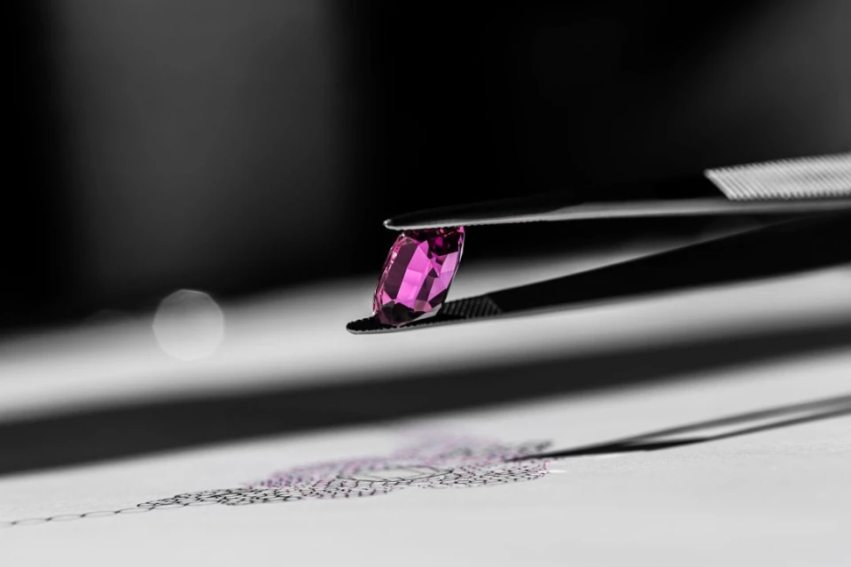 Close-up of pink gemstone held by metal tweezers, casting shadow on white surface against dark background with a circular light.