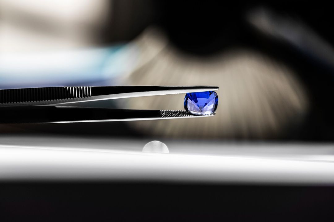 Close-up of vibrant blue gemstone held by tweezers against a blurred background, showcasing its cut and color.